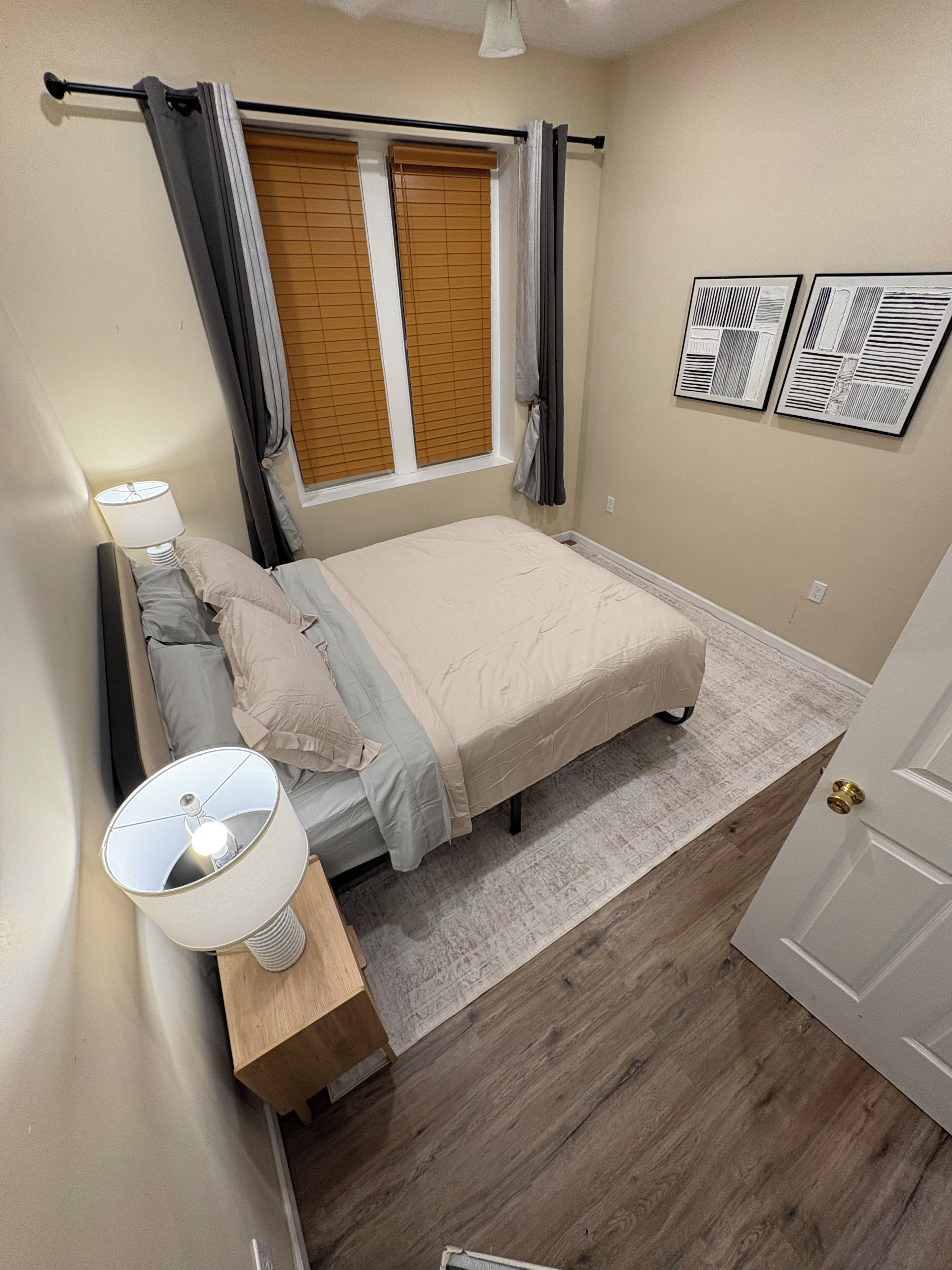 Un dormitorio con cama, mesita de noche, lámpara, ventana con persianas y dos rejillas de ventilación en la pared.