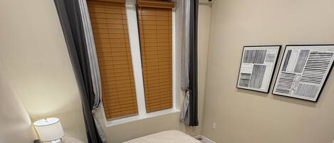 Un dormitorio con cama, mesita de noche, lámpara, ventana con persianas y dos rejillas de ventilación en la pared.