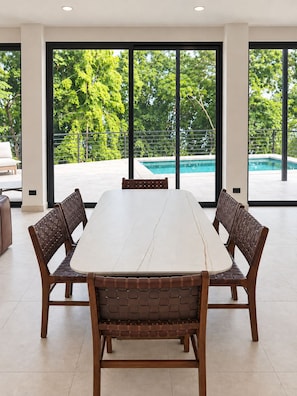 A spacious room with a large dining table and four chairs, large windows with a view of a swimming pool and trees. - Oceanview Villa · Private Pool Access · Panoramic Ocean Views (Garabito)