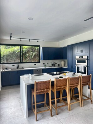 A modern kitchen with dark blue cabinets, a white countertop, and a central island with chairs. - Oceanview Villa · Private Pool Access · Panoramic Ocean Views (Garabito)