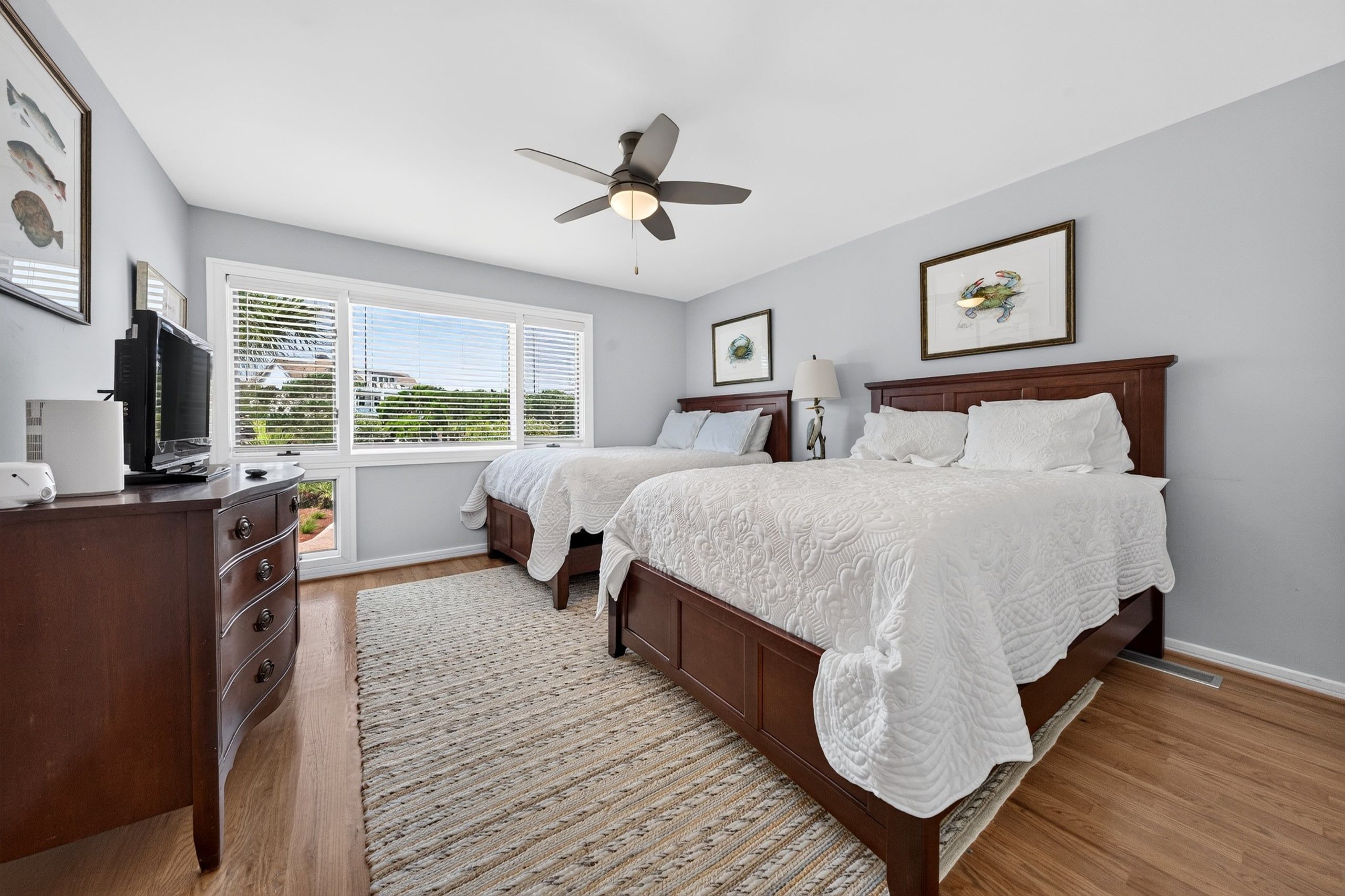 A bedroom with two beds, a ceiling fan, a TV, and artwork on the walls.