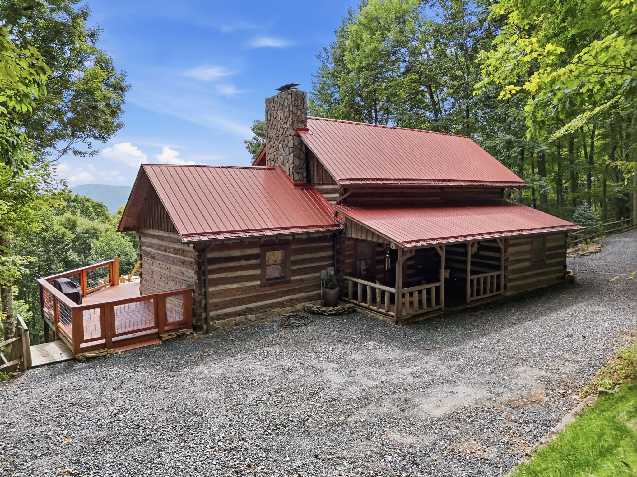A two-story log cabin with a red metal roof, a covered porch, and a gravel driveway.