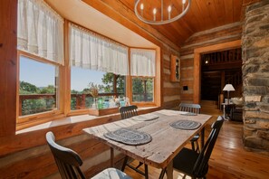 A wooden dining room with a large table and chairs, large windows with curtains, and a stone wall. - 2BR Mountain cabin with hot tub, game room, gas fireplace (West Jefferson)