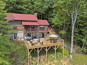 A log cabin with a red roof, a stone foundation, and a wooden deck with outdoor seating. - 2BR Mountain cabin with hot tub, game room, gas fireplace (West Jefferson)