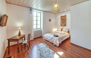 Une chambre avec un plancher en bois, un lit avec une literie blanche, un bureau avec une lampe, une chaise, un radiateur et une fenêtre avec des rideaux.