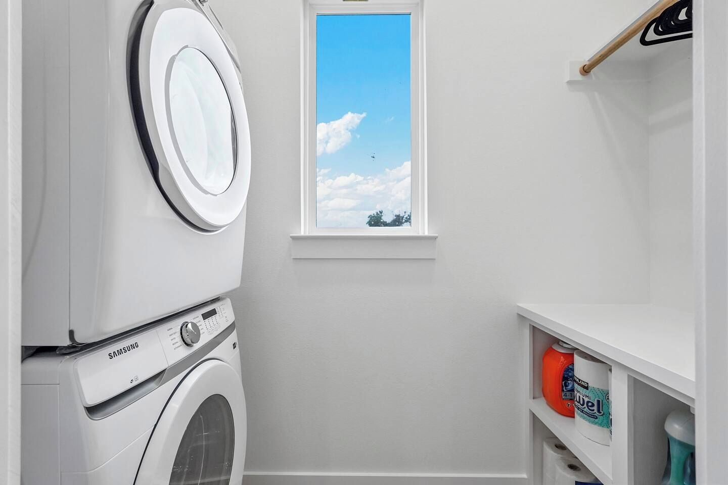 Uma lavanderia com máquina de lavar, secadora, janela com vista para o céu e prateleira com produtos de limpeza.