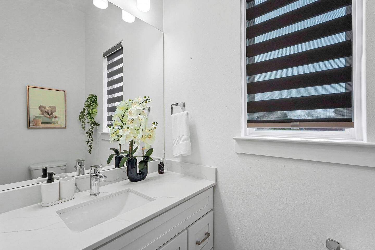 Une salle de bain moderne avec un lavabo blanc, un miroir et une fenêtre avec des stores.