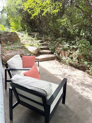 A patio with a black metal chair and cushions, surrounded by greenery and a stone pathway. - Red Rock Ridge- 2 Bedroom Suite (Wheat Ridge)