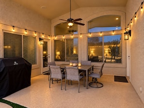 An outdoor patio with a table and chairs, a ceiling fan, string lights, and a grill.