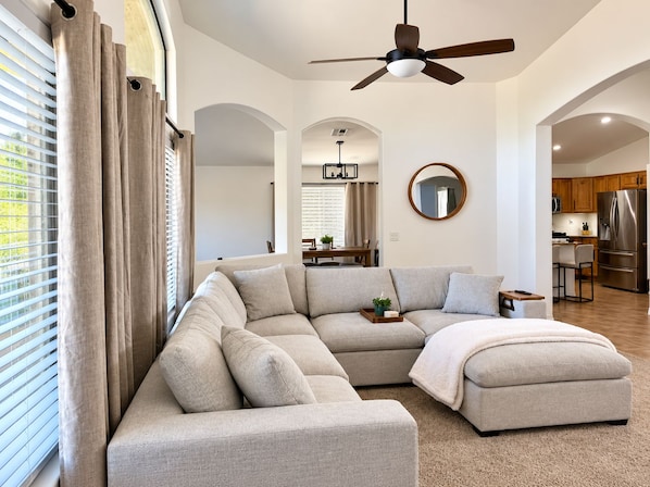 A modern living room with a sectional sofa, a ceiling fan, and a view into the kitchen.