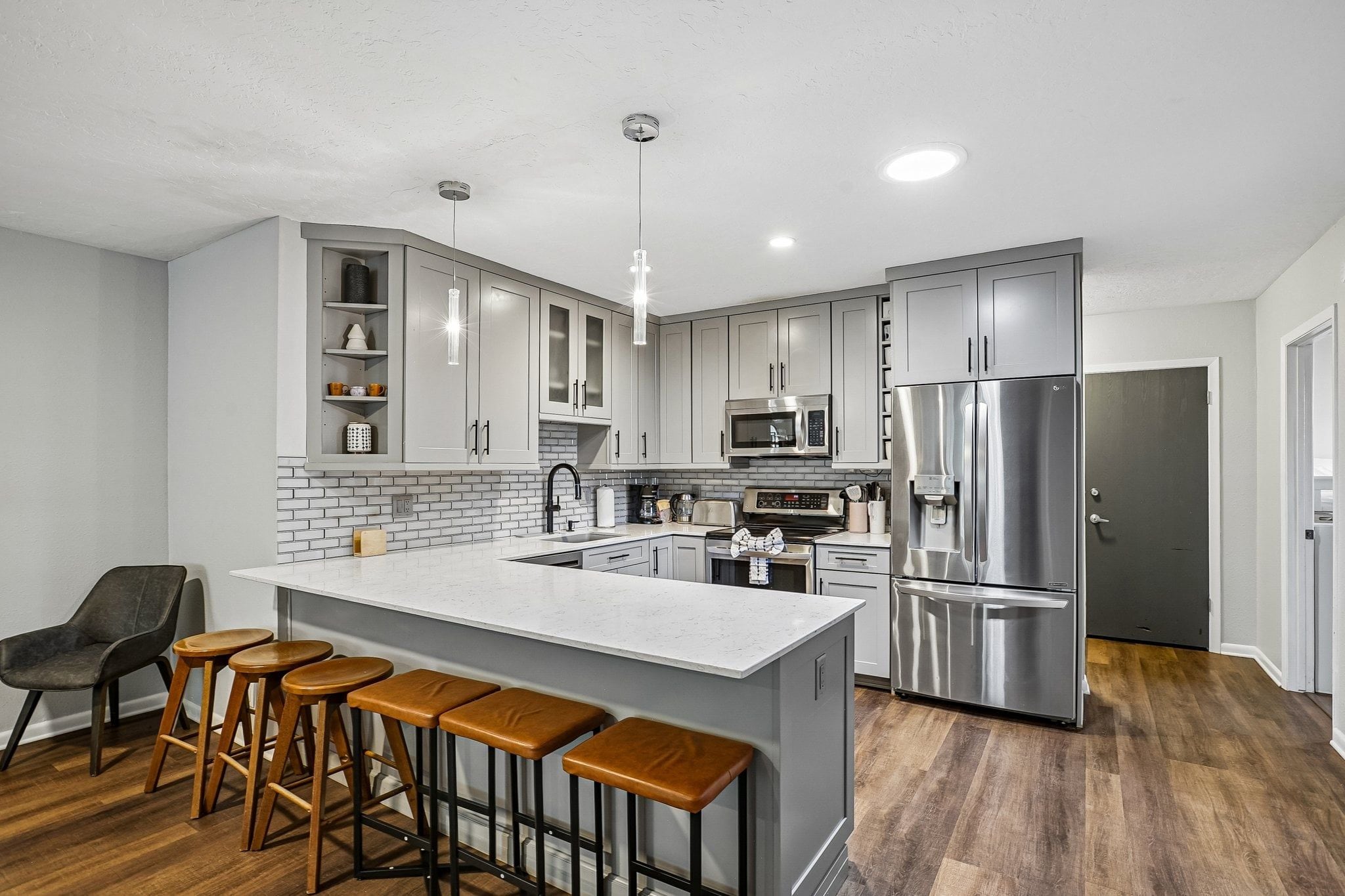 A modern kitchen with grey cabinets, a stainless steel refrigerator, a microwave, and a bar area with stools.