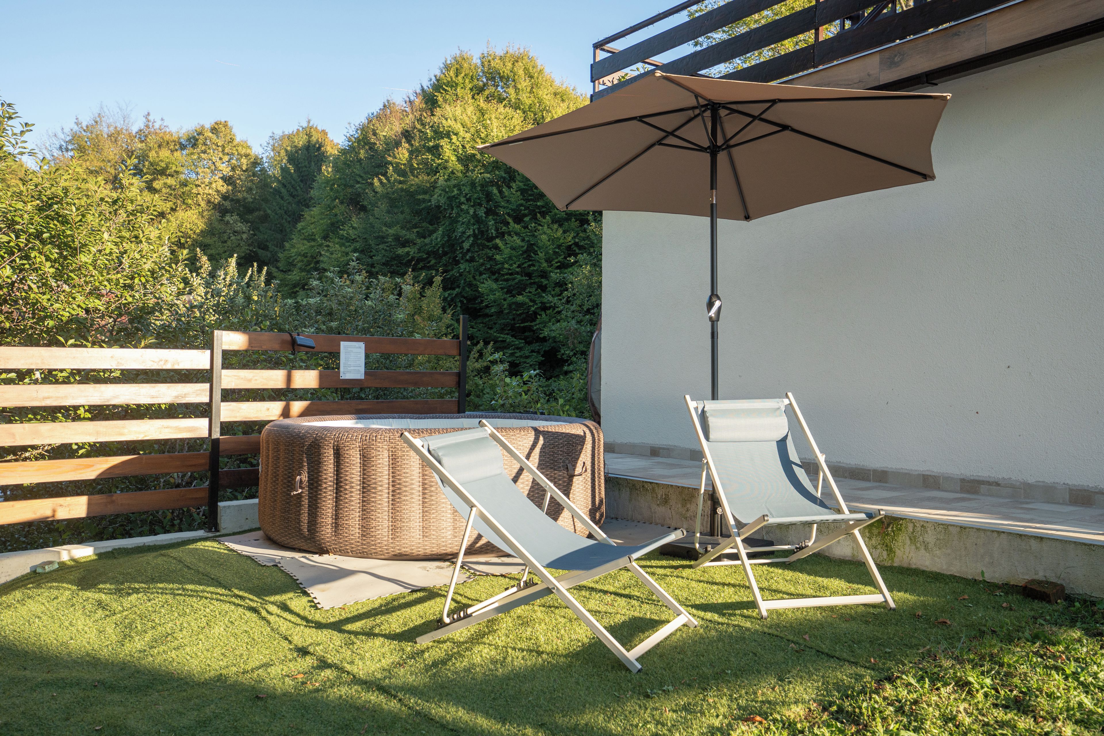 Un espace extérieur aménagé avec deux fauteuils confortables et un grand parasol, le tout adossé à une clôture en bois et à de la verdure.