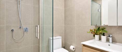 Un baño moderno con ducha envuelta en vidrio, inodoro blanco y tocador de madera con lavamanos y espejo.