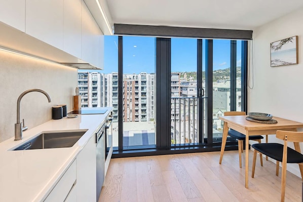 A modern kitchen with a large window, a dining area with a table and chairs, and a view of high-rise buildings. - Te Aro City Apartment - 1BR with Pool & Gym (Wellington)