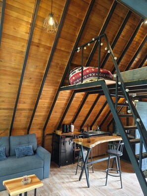 A loft-style room with a wooden ceiling, a bunk bed, a kitchenette, and a seating area.