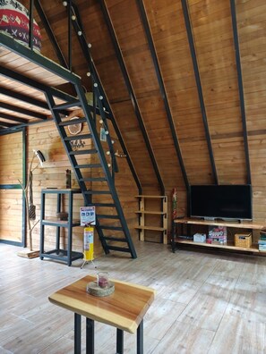 A loft-style room with a wooden floor, a TV on a shelf, a ladder, and a table with a glass. - Bird House Ecolodge (Barbosa)