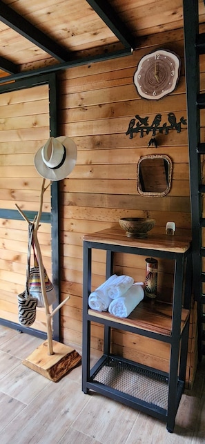 A room with wooden walls, a hat stand, a mirror, a clock, and a shelf with towels and a decorative bowl.