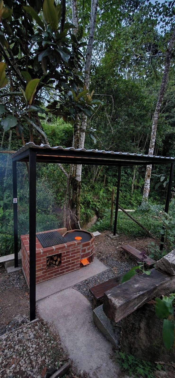 Ein Außenbereich mit Grillplatz aus Ziegelsteinen und einer Metallüberdachung, umgeben von Grünflächen und Bäumen.
