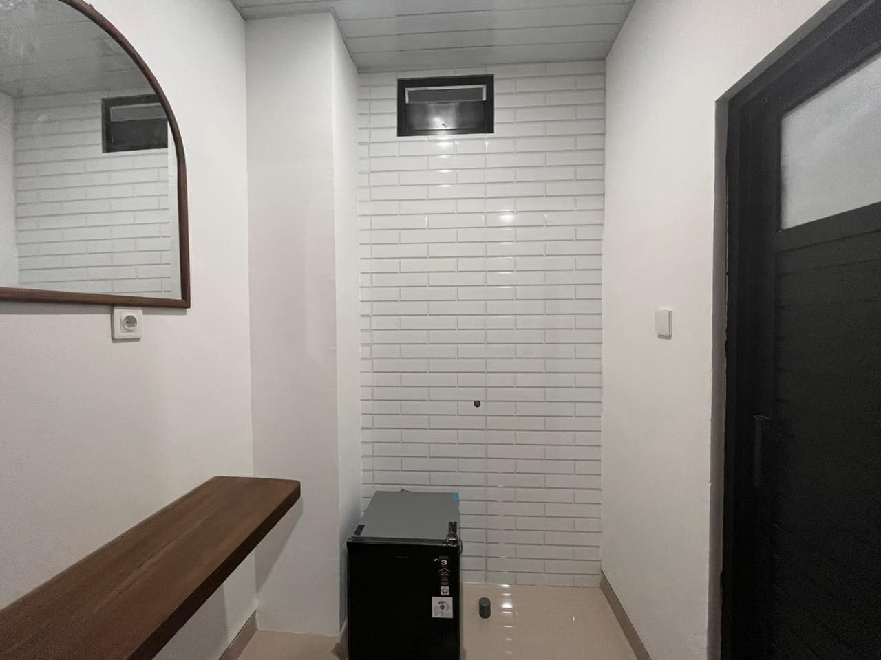 A hallway with white tiled walls, a wooden bench, a black storage unit, and a black door.