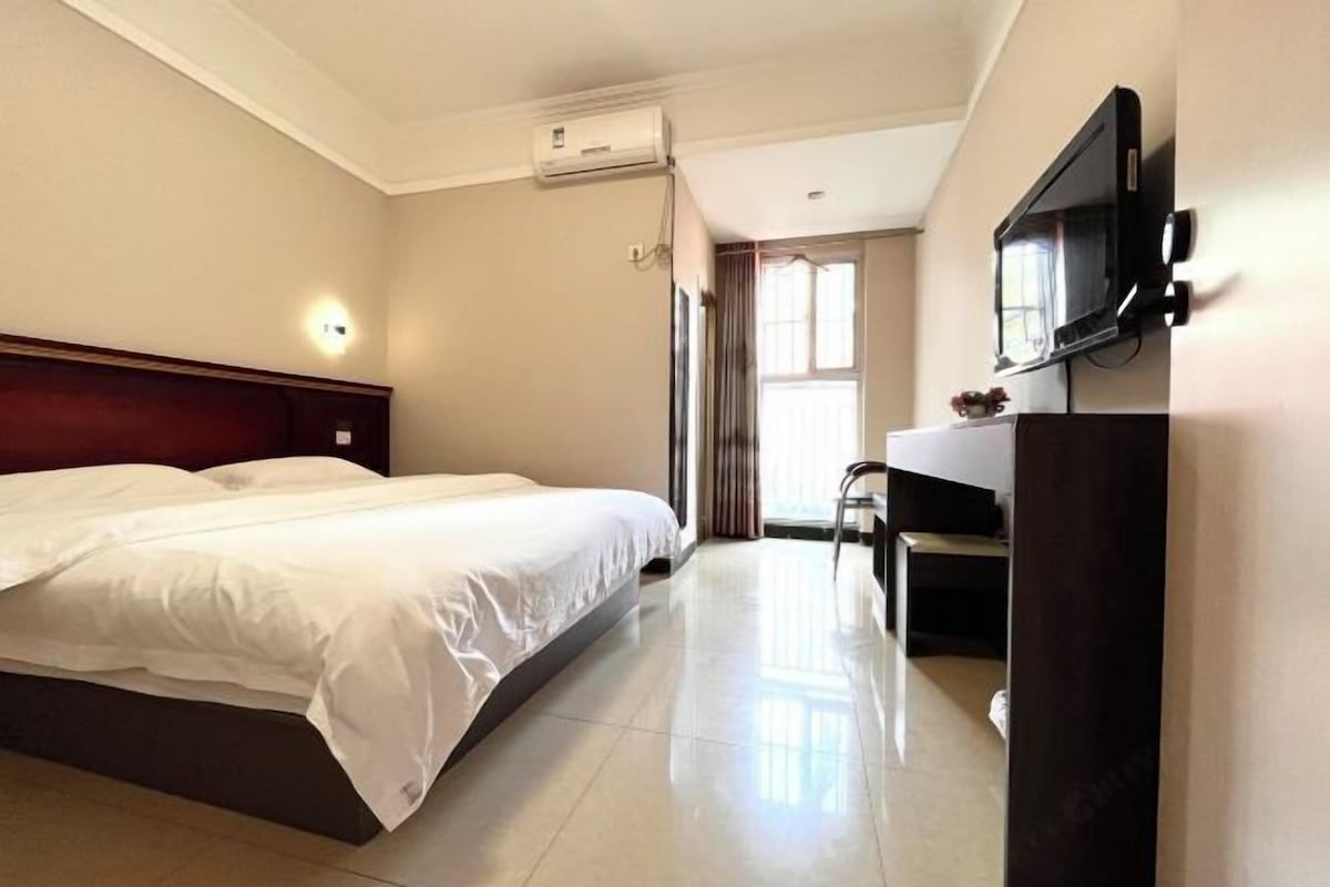 A hotel room with a bed, a desk, a chair, a TV, and an air conditioning unit.