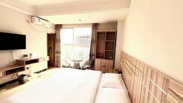A hotel room with a large bed, a TV mounted on the wall, a wooden wardrobe, and a window with curtains.
