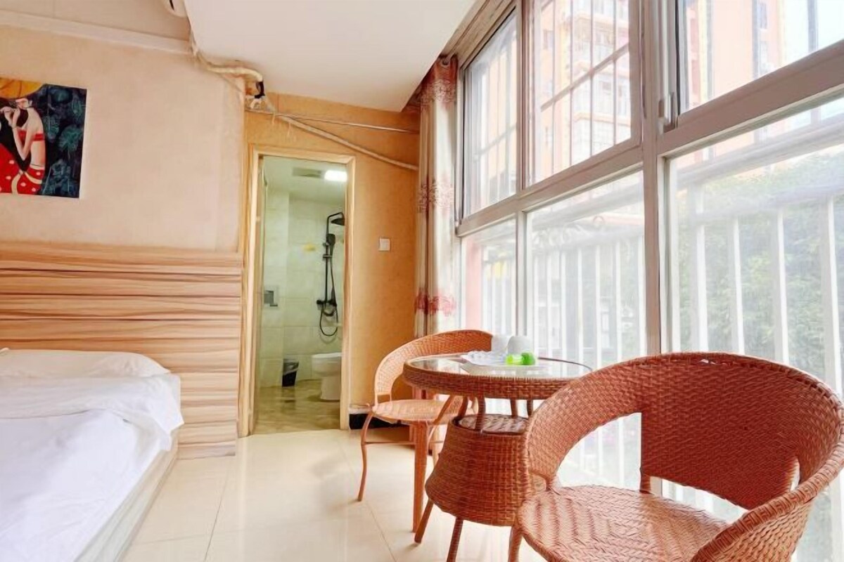 A hotel room with a bed, a dining table with chairs, a bathroom with a shower, and large windows.