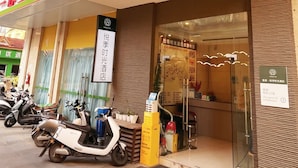 Un edificio con un letrero que indica 'MAXHILL INN' y una farmacia de salud. Hay scooters estacionados afuera, y un letrero que señala 'Habitaciones 3F Ascensor y Elevador'.
