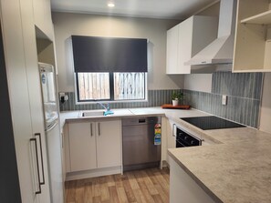 A compact kitchen with a built-in dishwasher, a window with a black roller blind, and a backsplash with vertical lines. - Fresh, Cosy & Handy to CBD, Lake, Waikato Hospital, Gym (2) and Dinsdale Shops (Hamilton)