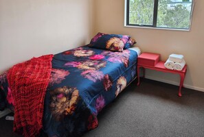 A single bed with a floral bedspread, a red throw blanket, and a matching pillow. There is a small red nightstand with a white alarm clock beside the bed. - Fresh, Cosy & Handy to CBD, Lake, Waikato Hospital, Gym and Dinsdale Shops (Hamilton)