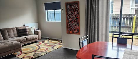 A living room with a sofa, a dining table with chairs, a patterned rug, an air conditioning unit, a window with curtains, and a wall-mounted artwork.