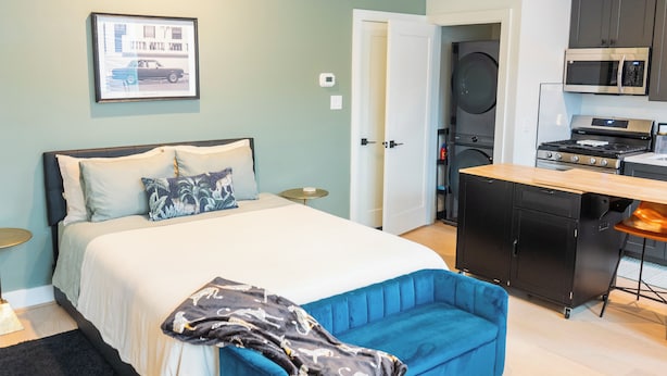 Ein Schlafzimmer mit einem Bett, Nachttischen, einem blauen Sessel und einer Küchenzeile inklusive Herd, Mikrowelle und Backofen.
