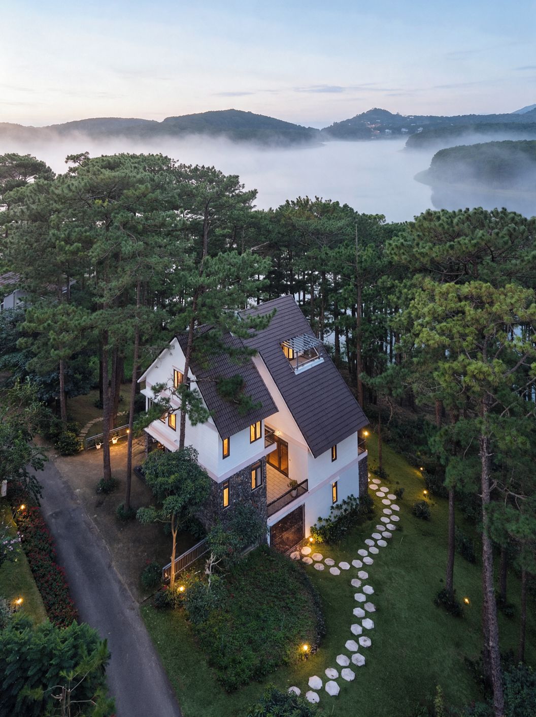 A modern two-story house with a sloped roof, surrounded by dense forest and a clear view of a lake.