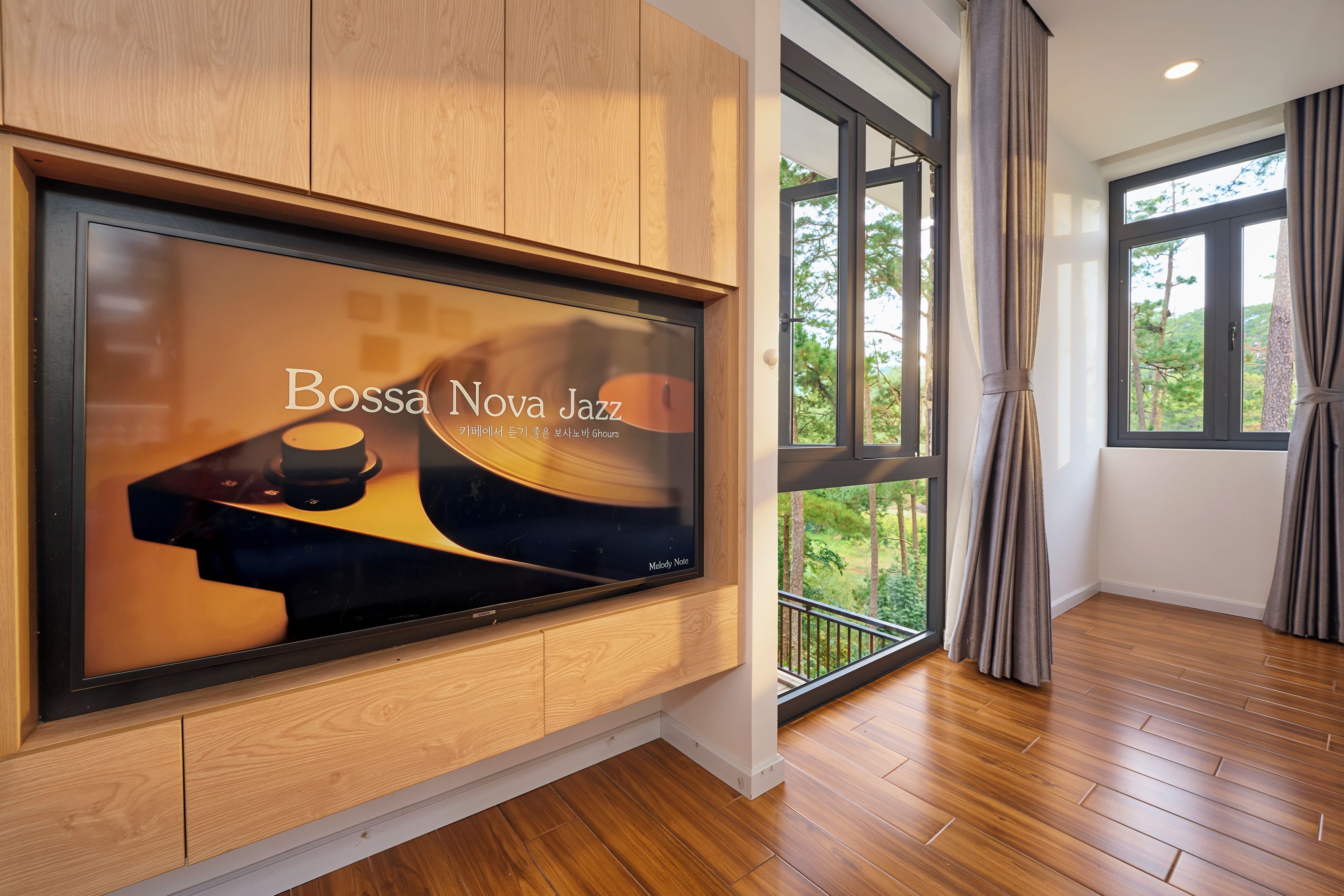 A modern interior with a large flat-screen TV mounted on a wooden wall, a balcony with a view of trees, and a wooden floor.