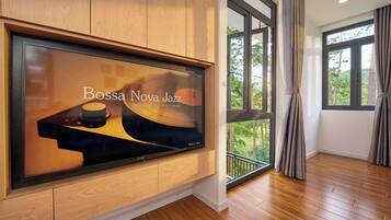 A modern interior with a large flat-screen TV mounted on a wooden wall, a balcony with a view of trees, and a wooden floor.