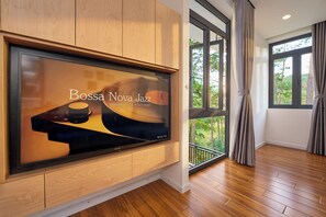 Un interior moderno con un televisor de pantalla plana grande montado en una pared de madera, un balcón con vistas a árboles y un suelo de madera.