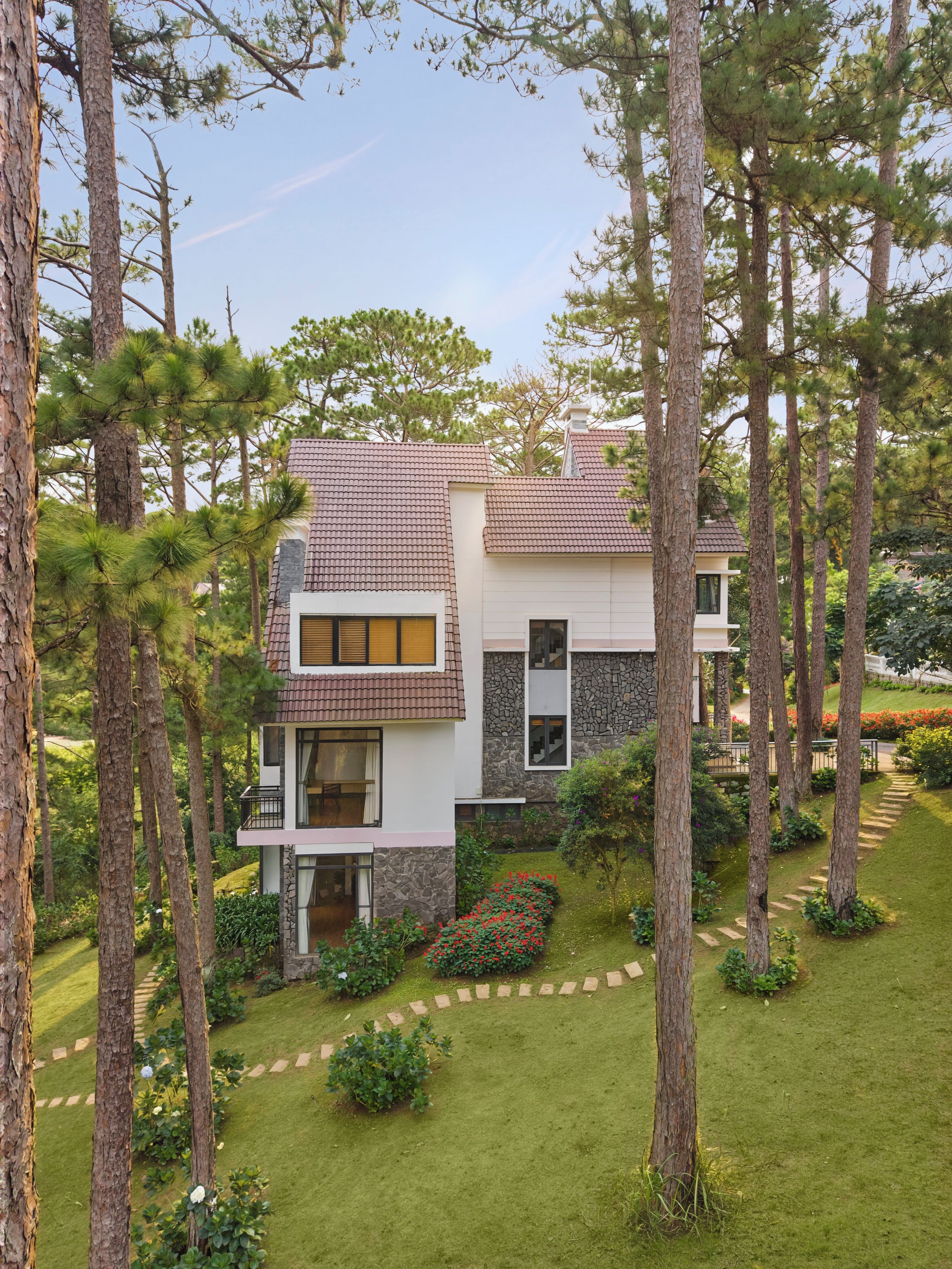A modern two-story house with a stone facade and a tiled roof, surrounded by a well-maintained garden and tall pine trees.