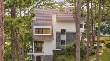 A modern two-story house with a stone facade and a tiled roof, surrounded by a well-maintained garden and tall pine trees.