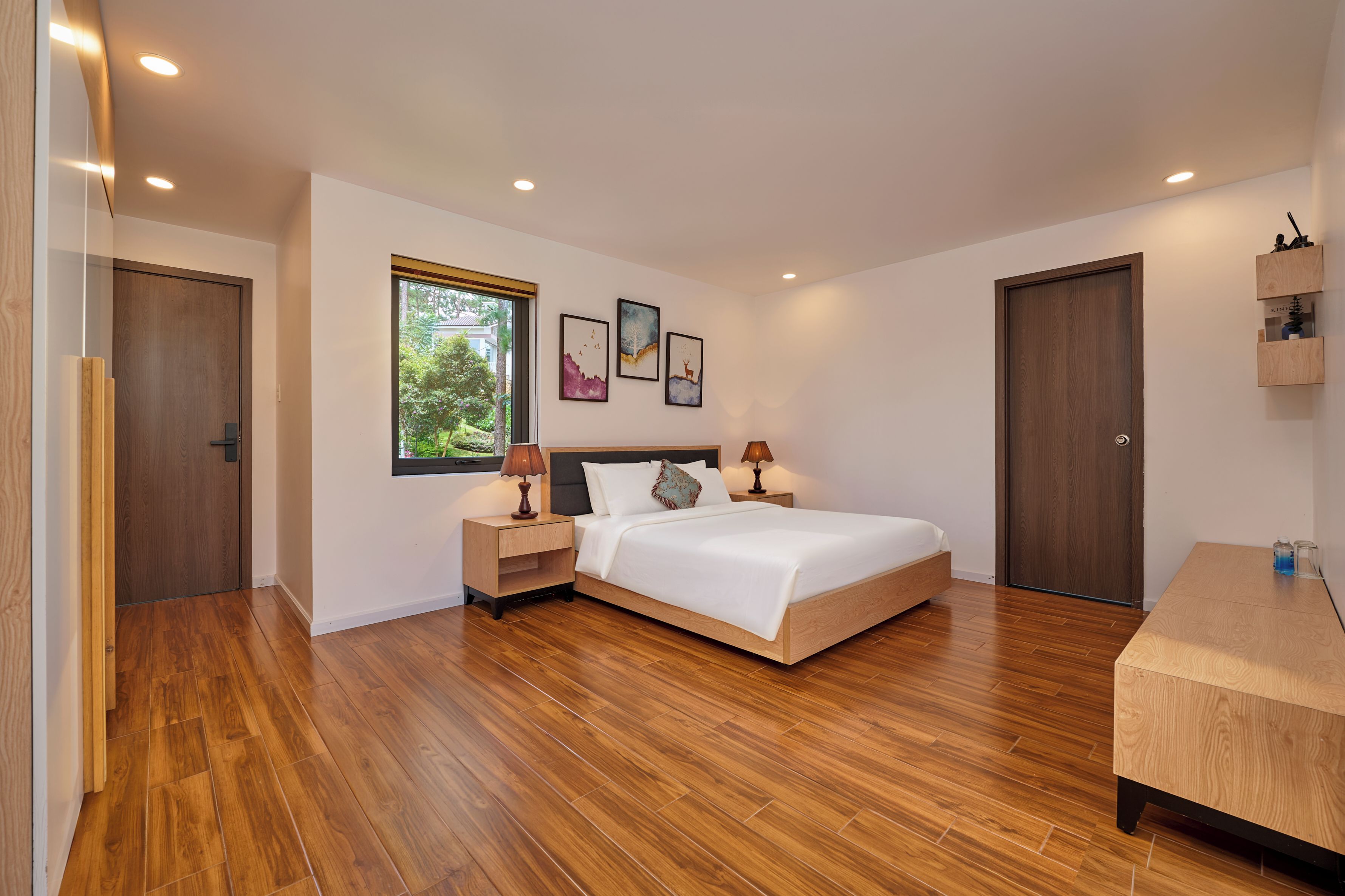 A modern bedroom with a wooden floor, a bed with white bedding, a nightstand with a lamp, and a window with a view of greenery.