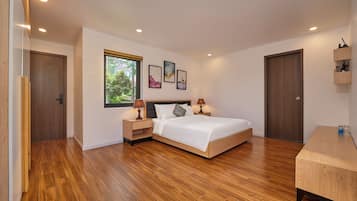 A modern bedroom with a wooden floor, a bed with white bedding, a nightstand with a lamp, and a window with a view of greenery.