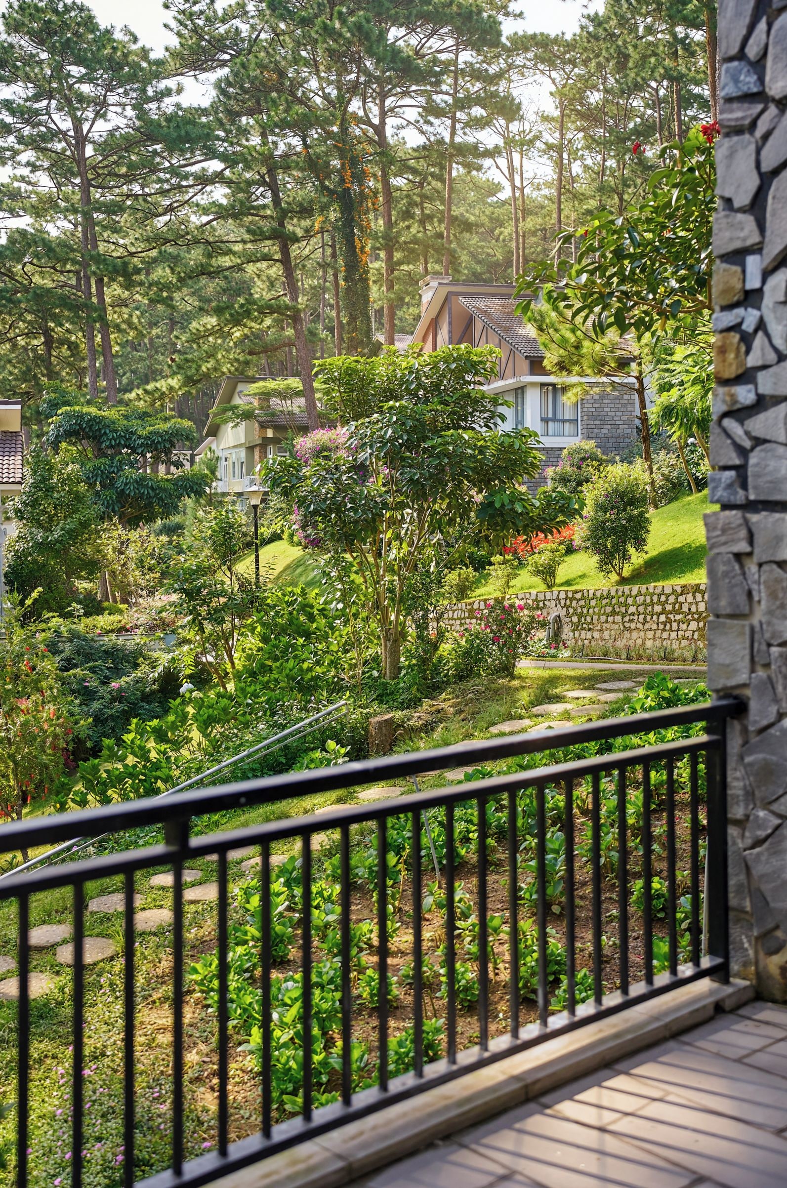 Un balcón con un muro de piedra, un jardín con diversas plantas y un pequeño estanque, y una casa tradicional al fondo.