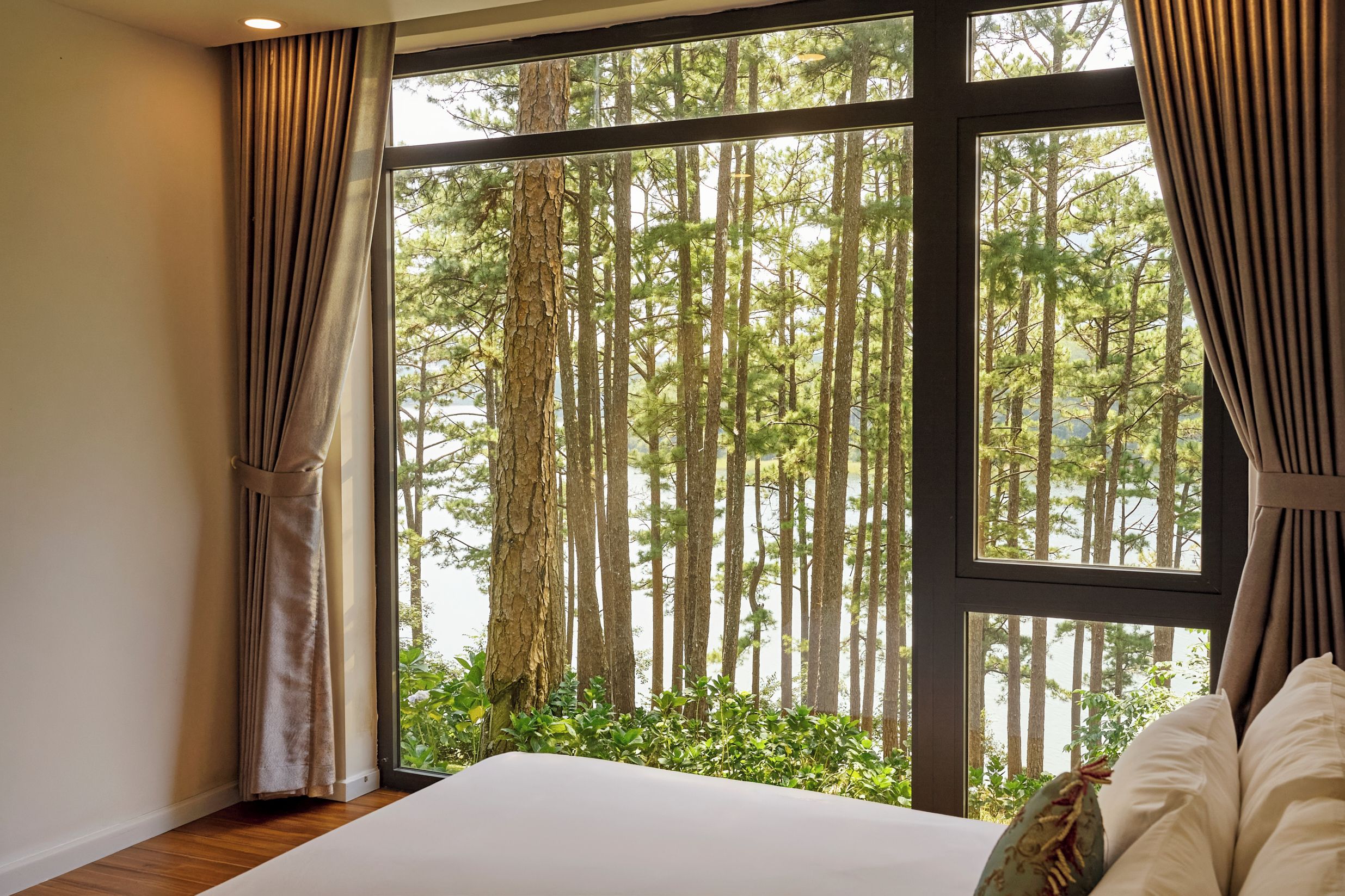 A bedroom with a large window, curtains, a bed, and a view of a forest.