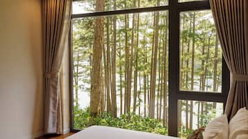 A bedroom with a large window, curtains, a bed, and a view of a forest.