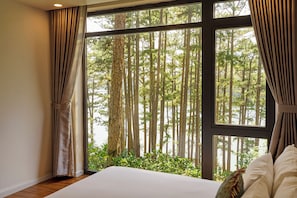 A bedroom with a large window, curtains, a bed, and a view of a forest.