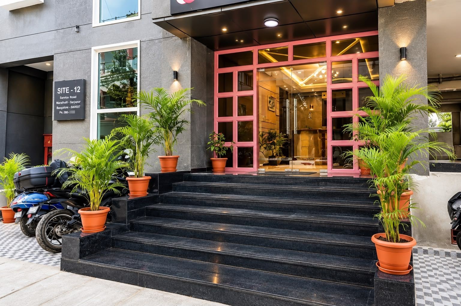 Entrada de un hotel con una escalera que conduce a una puerta de vidrio, rodeada de plantas en macetas y una moto estacionada.