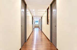 A long, narrow hallway with wooden flooring and doors on both sides, leading to a bright room at the end. - East Land Hotel Near Manyata Tech (Bengaluru)