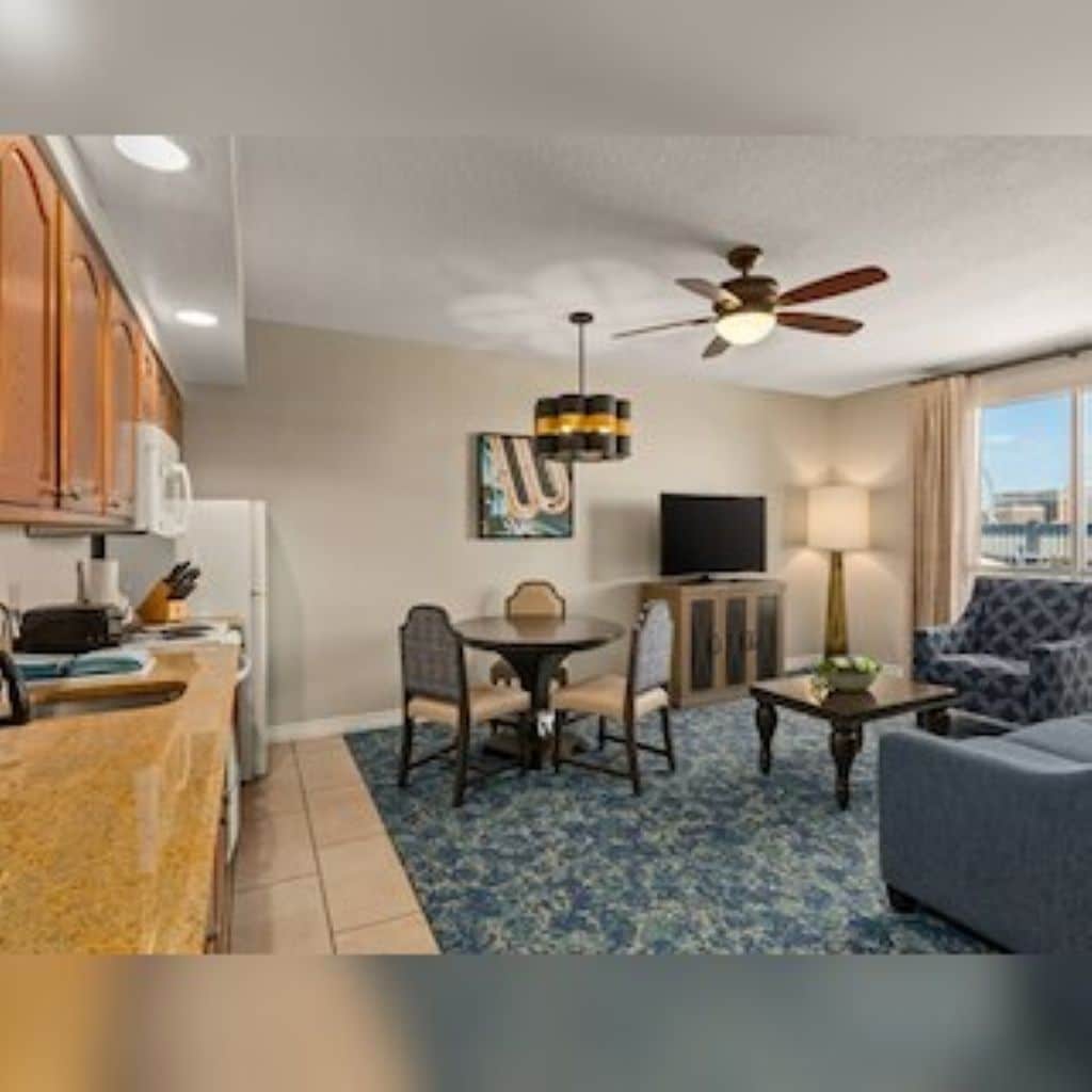 A hotel room with a kitchenette, a dining area with a table and chairs, a sofa, a TV, and a ceiling fan.