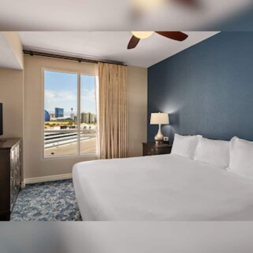 A hotel room with a large bed, a nightstand, a lamp, a ceiling fan, and a window with a view of the city.
