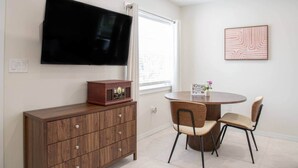 A room with a flat-screen TV mounted on the wall, a wooden cabinet with drawers, a round dining table with two chairs, and a framed picture on the wall. - #8 Beach Access King Studio (Sarasota County)