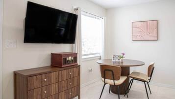 A room with a flat-screen TV mounted on the wall, a wooden cabinet with drawers, a round dining table with two chairs, and a framed picture on the wall.