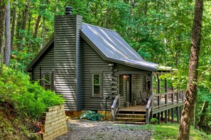 A log cabin with a porch and a chimney, surrounded by dense forest. - Piney Mountain Perch - Secluded Log Cabin with Firepit + Fireplace (Mineral Bluff)